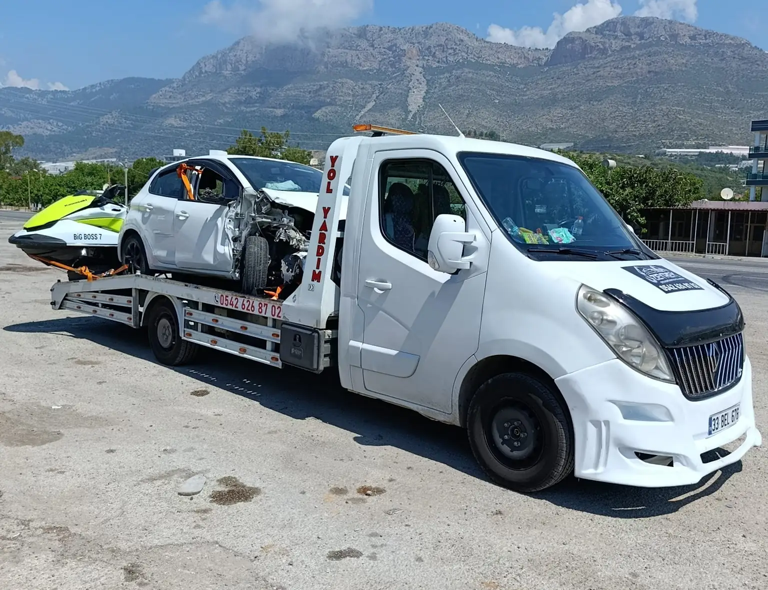 Döşemealtı için Zeyrek Oto Çekici’nin platformlu çekicisi, dağ manzarası önünde hasarlı bir otomobili ve bir jet ski’yi taşıyor