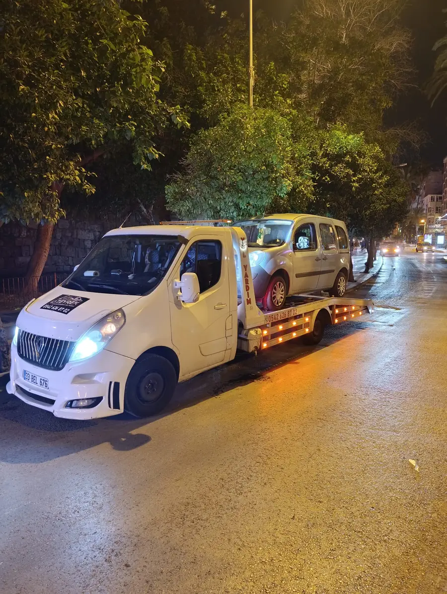 Akşam saatlerinde ışıkları yanan beyaz çekici, platformunda gümüş rengi bir hafif ticari araçla yol kenarında